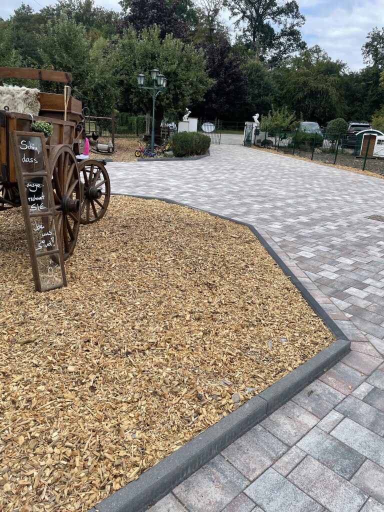 Das Bild zeigt eine großen Hof der gepflastert ist.in der Mitte ist eine Insel angelegt mit Rindenmulch und einer alten Kutsche